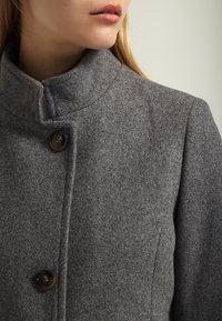 Grå ullrock med hög krage, tre knappar fram och en mjuk textur. Har en skräddarsydd design och neutral färg.