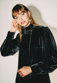 Abito in velluto nero con collo alto e dettagli con bottoni. Maniche lunghe e gonfie. Grandi orecchini dorati e colorati. Mano adornata con un anello vistoso.