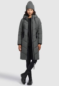 Donna che indossa un lungo cappotto imbottito grigio con cappuccio, un berretto di maglia grigio, pantaloni neri e stivali neri, in piedi con le gambe incrociate su uno sfondo bianco.