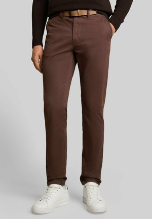 Mężczyzna noszący brązowe spodnie slim fit z beżowym paskiem, ciemny sweter, białe trampki, stoi z jedną ręką w kieszeni.