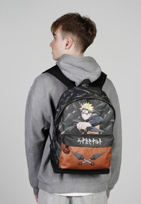 Schwarzer Rucksack mit Naruto-Design, das eine Figur zeigt, die Shurikens wirft. Orangefarbener unterer Abschnitt, verstellbare Riemen und Reißverschlusstasche.