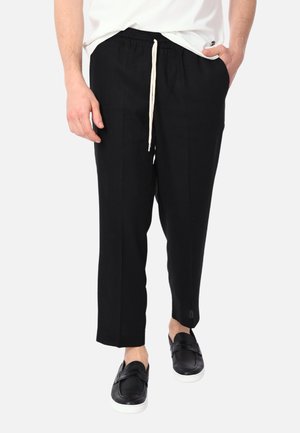 Uomo che indossa pantaloni neri con coulisse e scarpe slip-on nere, una mano in tasca, in piedi contro uno sfondo bianco.