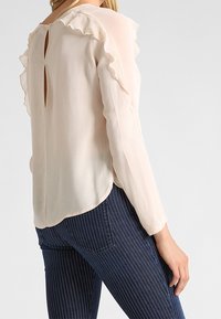 Blusa beige chiaro con spalle arricciate, dettaglio a occhiello sul retro e maniche lunghe; abbinata a jeans scuri a righe. Tessuto liscio e leggero.