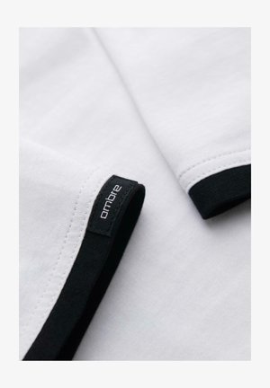 Tissu blanc avec des bordures noires et une étiquette de la marque "ombre" cousue près de l'ourlet sur un vêtement plié.