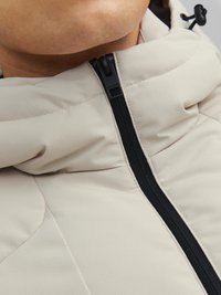 Beige gepolsterte Jacke mit hohem Kragen, schwarzem Reißverschluss und strukturiertem Stoff. Sie weist ein modernes Design mit ausgeprägten Steppdetails auf.