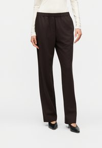 Γυναίκα που φοράει καφέ σκούρο, ψηλόμεσο παντελόνι με ίσια μπατζάκια και ελαστική ζώνη μέσης, σε συνδυασμό με μαύρες μυτερές γόβες, στέκεται μπροστά σε λευκό φόντο.