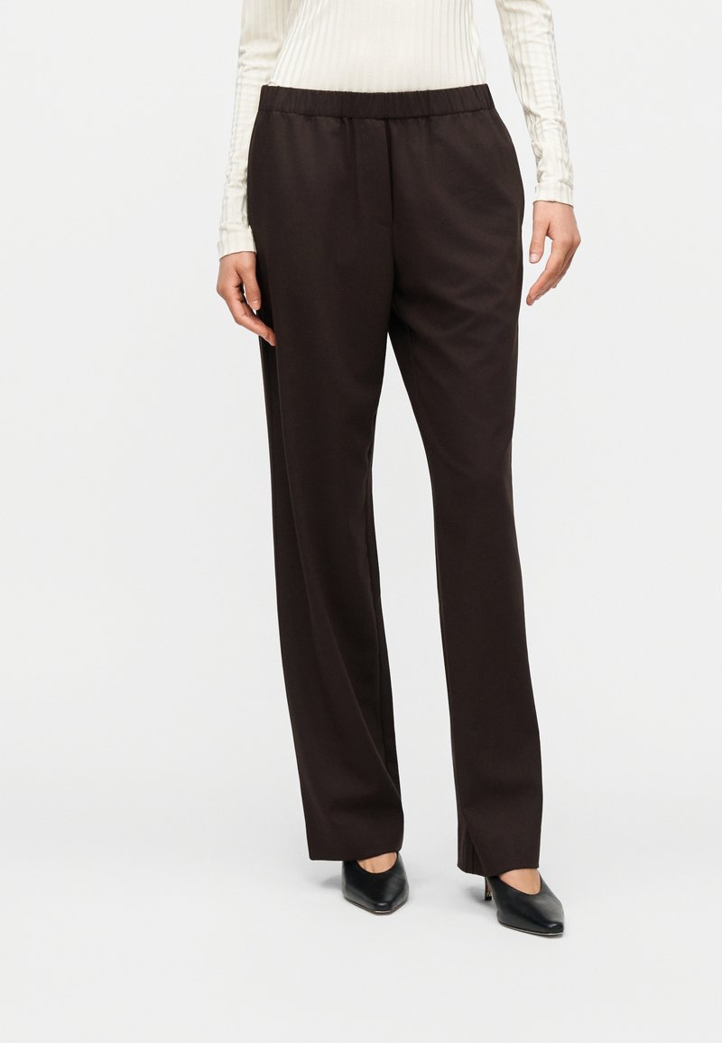 Γυναίκα που φοράει καφέ σκούρο, ψηλόμεσο παντελόνι με ίσια μπατζάκια και ελαστική ζώνη μέσης, σε συνδυασμό με μαύρες μυτερές γόβες, στέκεται μπροστά σε λευκό φόντο.