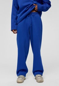 Blauwe sweatpants van zacht materiaal, met een losse pasvorm, elastische tailleband met trekkoord en zijzakken. Gecombineerd met grijze sneakers.
