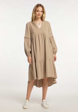 Frau trägt ein lockeres beiges langärmliges Kleid mit V-Ausschnitt und weißen Sneakers, steht vor einem schlichten weißen Hintergrund.