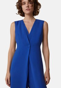 Salopette blu senza maniche con scollatura a V profonda, chiusura frontale con bottoni e design drappeggiato. Il tessuto liscio e la vestibilità sartoriale esaltano la silhouette.