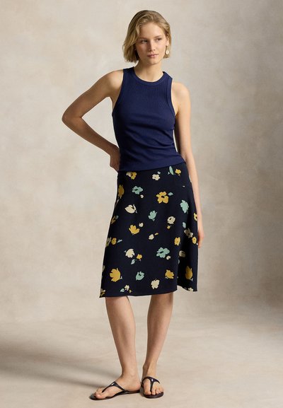 Jeune femme vêtue d'un haut sans manches bleu marine, d'une jupe midi florale foncée, des sandales, debout avec une main sur la hanche sur un fond uni.