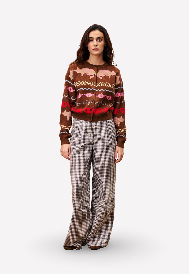 Bruine gebreide cardigan met bloem- en golfpatronen, gecombineerd met wijde, geruite broeken in neutrale tinten. Model staat rechtop.