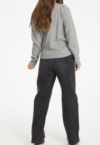 Femme aux longs cheveux bruns portant un pull gris clair et un jean ample gris foncé, debout de dos sur un fond blanc.