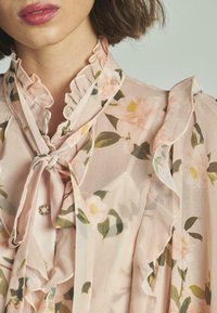 Blusa floral en tejido ligero y translúcido de color rosa. Presenta un cuello con volantes, un lazo de cinta y botones con un patrón floral y hojas verdes.