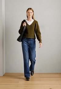 Oliven grønn strikket genser med V-hals, hvit lagdelt topp under, kombinert med løse blåjeans og svarte sko, bærende en svart veske.