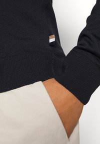 Suéter navy con puños y dobladillo acanalados. Presenta una pequeña etiqueta con franjas en marrón, blanco y negro en el lateral. Fabricado con una tela suave.