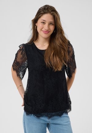 Jeune femme aux longs cheveux bruns portant un haut en dentelle noire avec des manches transparentes et un jean bleu clair, souriante, les mains derrière le dos.