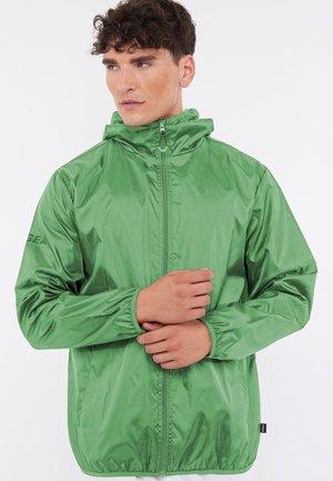 Jeune homme portant une veste à capuche zippée vert brillant, debout avec les mains jointes à la taille, regardant légèrement sur le côté.