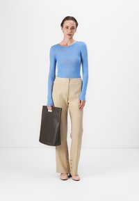 Blaues Langarmoberteil, cremefarbene plissierte Hose mit seitlichen Knöpfen, hellrosa Schuhe, hält eine dunkelbraune Tragetasche. Einfarbiger Hintergrund.