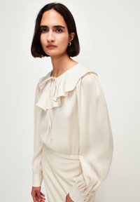 Cremefarvet bluse med volangkrave, lange ærmer og en bindedetalje ved halsen. Stoffet har en blød tekstur og en subtil glans.