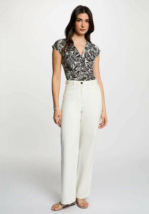 Donna con top a stampa di foglie bianco e nero, pantaloni bianchi a gamba larga a vita alta e sandali con cinturini, in piedi davanti a uno sfondo semplice.