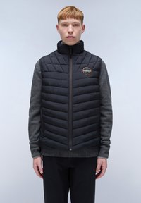 Schwarze Steppweste mit hohem Kragen, ausgestattet mit gesteppter Textur und einem grauen Pullover darunter. Enthält ein rundes Logo auf der Brust.