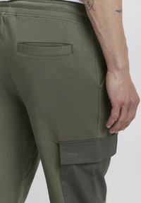 Olive green sweatpants with a smooth texture, featuring a side pocket in a darker shade and a back pocket above the waistband.