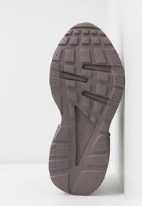 Semelle de chaussure en caoutchouc brun foncé avec un motif de bande de roulement texturé, présentant des rainures et des crêtes pour l'adhérence. Forme incurvée avec une paroi latérale.