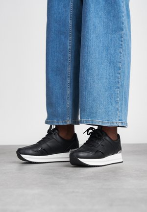 Baskets en cuir noir avec semelles blanches, présentant un design épuré, un système de laçage et de subtils accents latéraux. Associées à un jean large bleu clair.