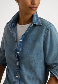 Gaiši zils džinsa krekls ar pogājamu apkakli, teksturētu audumu un baltām pogām, uzvilkts virs balta T-krekla.