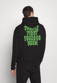 Man med ryggen mot kameran iförd en svart hoodie med neongrön text "FAMILY FIRST ICEBERG CREW" på ryggen framför en enfärgad bakgrund.