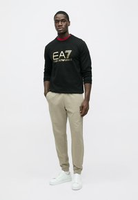 Schwarzes Sweatshirt mit goldenem Logo, beige Jogginghosen und weiße Turnschuhe. Glatte Texturen und lockere Passform. Einfaches Design ohne Muster.