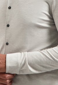 Helles grau Langarmhemd aus strukturiertem Stoff, mit schwarzen Knöpfen und sichtbar ausgearbeitetem Manschettenbereich. Nahaufnahme des Ärmels und der Knopfleiste.