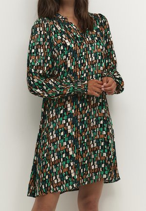 Mujer con vestido hasta la rodilla con mangas largas abullonadas y patrón abstracto de bloques en verde, marrón, blanco y negro.