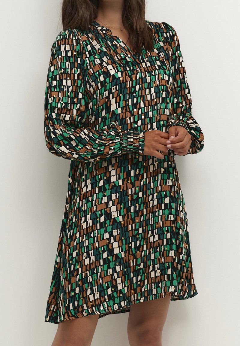 Nainen, joka pukeutunut polvipituiseen mekkoon, jossa on muhkeat pitkät hihat ja abstrakti vihreän, ruskean, valkoisen ja mustan ruutukuosi.