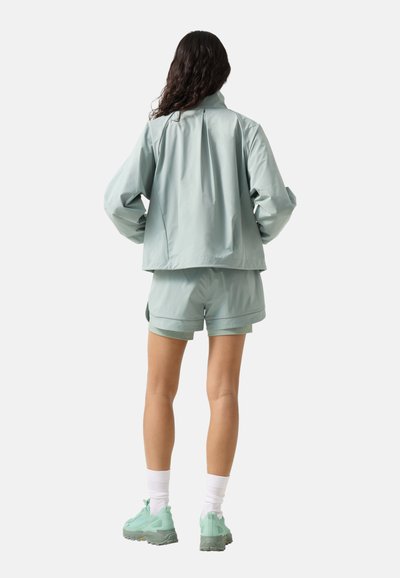 Kvinde med langt mørkt hår iført lysegrøn jakke, matchende shorts, hvide sokker og grønne sneakers, stående med hænderne på hofterne, set bagfra.