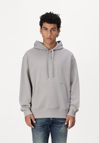 Helles grauer Hoodie mit einer Fronttasche, Zugbändchen und gesticktem Logo. Das Material scheint weich zu sein, mit gerippten Bündchen und Saum. Jeans sind darunter sichtbar.