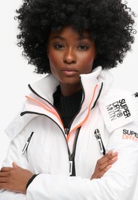 Biała kurtka puchowa z czarnymi i pomarańczowymi zamkami, z kieszeniami na zamek i markowymi akcentami. Teksturowany materiał, wysoki kołnierz.