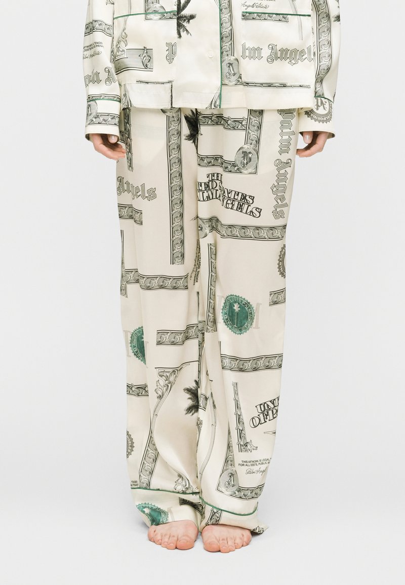 Άτομο που φοράει κρεμ πιζάμα με μαύρο και πράσινο σχέδιο, όρθιο ξυπόλητο μπροστά σε απλό ανοιχτό φόντο.