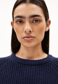 Marineblauw gebreid sweater met een geribbelde textuur en ronde halslijn. Close-up focus op gezicht met steil haar en natuurlijke make-up.