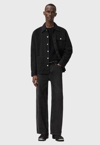 Czarna kurtka z płótna z frontową kieszenią, zestawiona z czarnymi szerokimi dżinsami. Model ma na sobie czarne buty oraz teksturowany sweter. W widocznych miejscach znajduje się srebrne guziki.