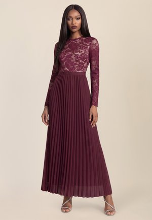 Kvinde i en vinrød blonde-top med lange ærmer og med en plisseret nederdel, stående mod en ensfarvet beige baggrund.