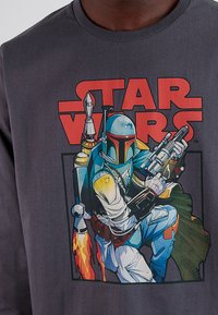Camiseta gris con una ilustración colorida de Boba Fett, con el texto "Star Wars" en negrita arriba. El diseño incluye detalles intrincados y colores vibrantes.