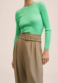 Grünes geripptes Strickoberteil mit langen Ärmeln, kombiniert mit hochgeschnittenen, beigen Faltenhosen, die über einen Gürtel mit Schnalle verfügen.