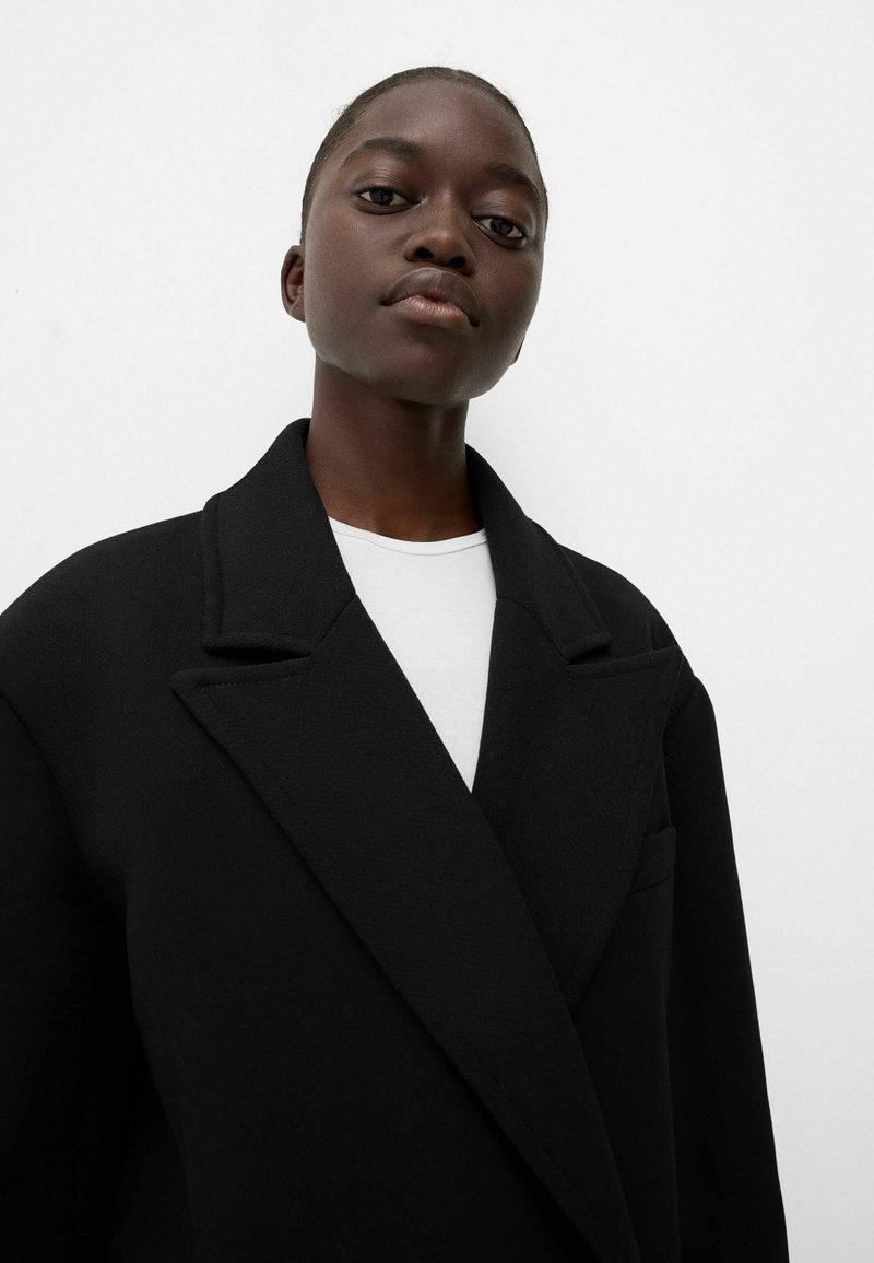 Schwarzer Wollmantel mit Reverskragen, mit strukturiertem Finish und einer Brusttasche. Über einem weißen Hemd getragen, vor einem neutralen Hintergrund.