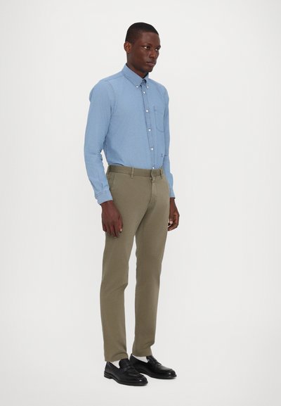 Chemise boutonnée bleu clair avec une poche poitrine, associée à un pantalon khaki. Modèle portant des chaussures noires, se tenant sur un fond blanc uni.