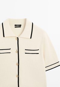 Polo di maglia color crema con bordini neri. Presenta un colletto classico, quattro bottoni e due tasche orizzontali decorative. Tessuto testurizzato.