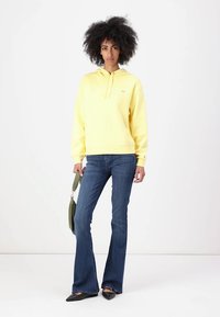 Felpa gialla con cappuccio e tasca a marsupio, abbinata a jeans blu svasati. Il modello tiene una borsa verde, mostrando un look casual.