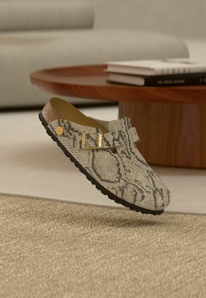 Slip-on sko med slangeprint, guldlås og tekstureret sål placeret på et tæppe nær et træ sofabord med stablede bøger.