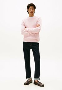 Maglione rosa chiaro in maglia a treccia con colletto a coste, abbinato a jeans blu scuro e mocassini marroni. Il modello è in piedi con le braccia incrociate.